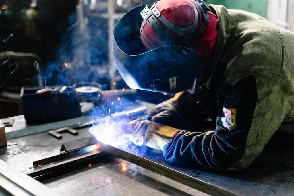 Welder wearing protective gear fusing metal components on a fabrication table with visible sparks and welding arc.